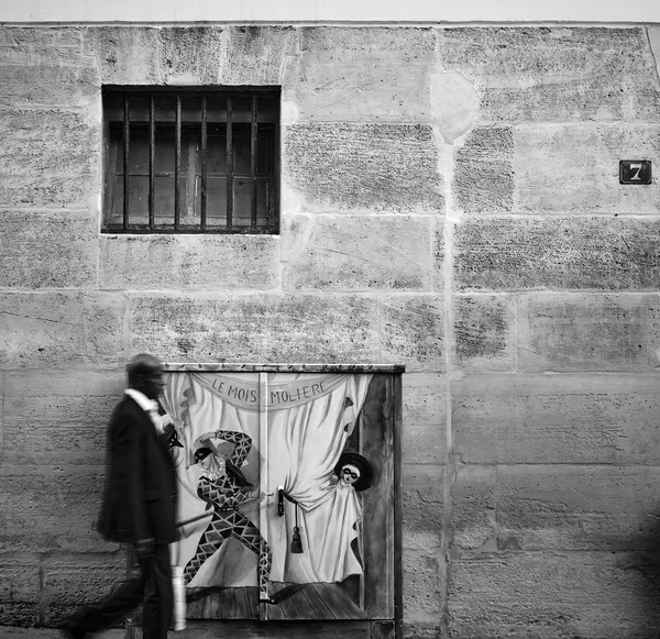 A man walks past a painted Molière theater panel set against an ancient stone wall, street photography with theatrical overtones in Versailles