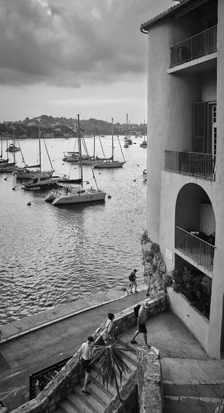 Sailboats moored in the harbor of Villefranche-sur-mer under gathering storm clouds, figures descending stone steps along the waterfront buildings