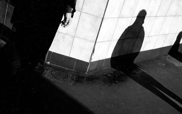 Dramatic shadows and silhouettes play across a Parisian sidewalk, a hand and human forms dissolving into stark contrasts of light and dark