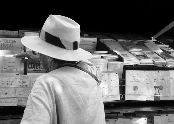 A man in a wide-brimmed hat browses a Parisian bookstall, his silhouette framed by rows of literary treasures in this elegant black and white street portrait