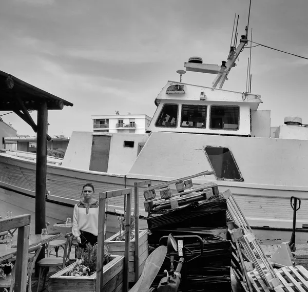A woman stands amid fishing boats and stacked crates in the port of Sète, a portrait of resilience against the rough texture of harbor life