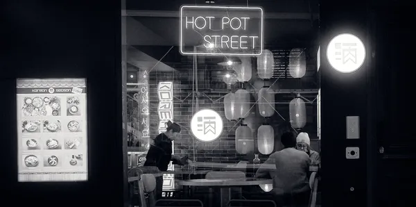 Diners glimpsed through the glowing window of a Parisian Asian restaurant at night, paper lanterns and neon signs painting an intimate nocturnal scene