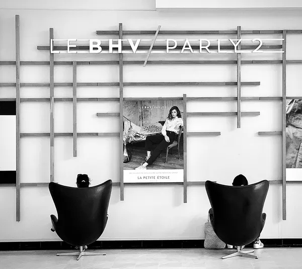 Two figures sink into oversized designer chairs beneath a department store advertisement, an ironic scene of consumer culture and disconnection