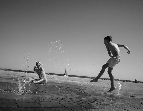 Children splash and play on a sun-drenched waterfront in Sète, frozen droplets of water suspended mid-air in this joyful black and white summer scene