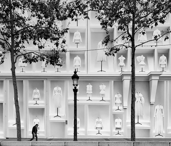 A lone passerby dwarfed by a monumental luxury fashion display, white garments on mannequins towering above the Parisian sidewalk like a temple of couture