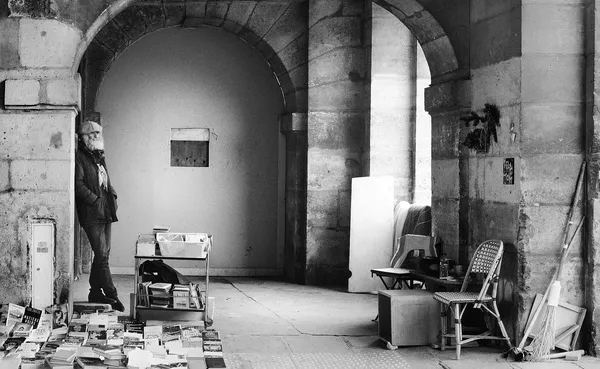 A bookseller leans against a stone archway surrounded by stacks of old books, a philosopher-like figure holding court in a Parisian passage