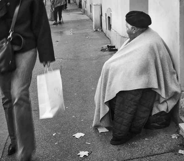 A person wrapped in a blanket sits on a Versailles sidewalk as a shopper strides past, two worlds colliding in a stark black and white street photograph