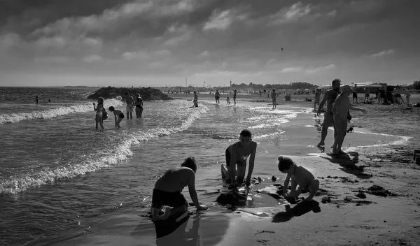 Beachgoers scatter along the shore under a dramatic cloudy sky, waves catching the light in this black and white seaside scene from Sète