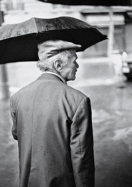 An elegant elderly gentleman shelters under an umbrella on a rainy Parisian day, his flat cap and suit jacket evoking a bygone era of quiet dignity