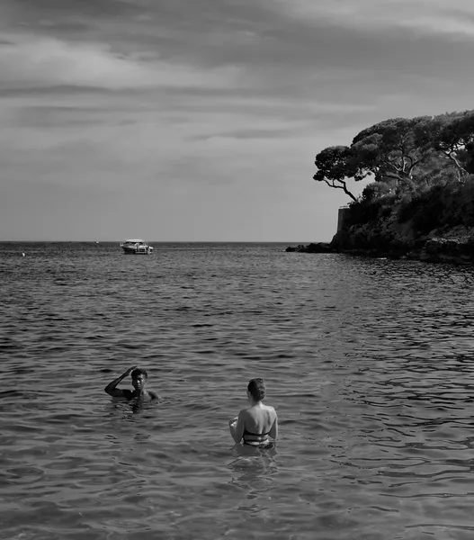 Two swimmers wade in the calm Mediterranean waters off Saint-Jean Cap Ferrat, pine trees and a distant boat completing a serene Riviera scene