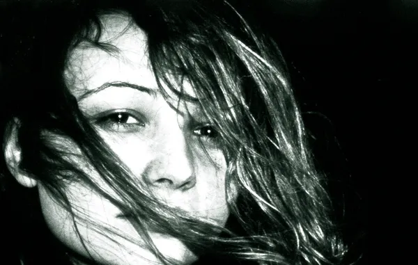 A woman's face emerges from darkness, windswept hair veiling her gaze in this intimate close-up portrait charged with mystery and sensuality