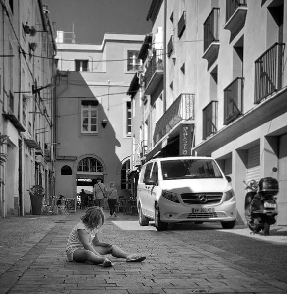 A young girl sits alone on the sunlit cobblestones of a Sète street, absorbed in her own world while the town carries on around her