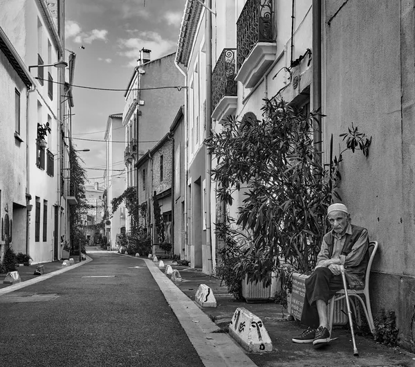 An elderly man sits with his cane on a quiet street in Sète, a sentinel of time watching over the empty Mediterranean alleyway
