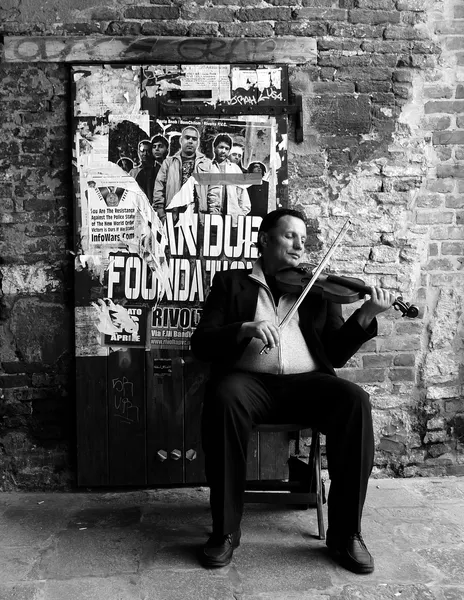 A street violinist plays seated against a weathered brick wall covered in torn posters in Venice, classical music defying the urban decay