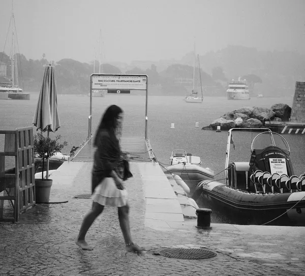 A woman strides past the harbor of Villefranche-sur-mer in soft motion blur, her silhouette dissolving against the misty Mediterranean backdrop