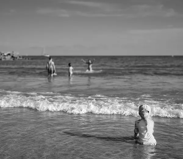 A small child sits alone in the shallow waves of a Sète beach, watching others play in the distance, a quiet moment of childhood contemplation by the sea