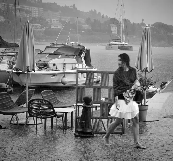 A mother and child walk along the rainy quay of Villefranche-sur-mer, motion blur lending an ethereal quality to their figures against moored boats and hazy hills
