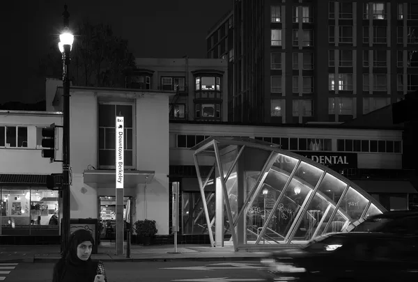 A woman stands alone near the glowing glass canopy of Downtown Berkeley station at night, urban solitude beneath the city lights