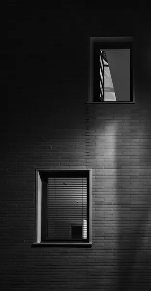 Two windows glow on a dark brick wall in Versailles at night, blinds and reflections creating an intimate study of light, shadow, and urban solitude