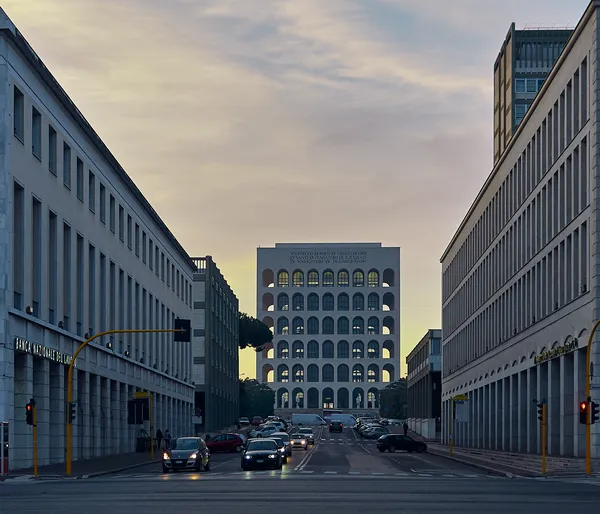 The Palazzo della Civiltà Italiana stands centered at the end of a Roman boulevard at dusk, its arched facade glowing amber against a pastel sky