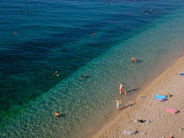 Aerial view of swimmers and sunbathers on a Dubrovnik beach, turquoise waters graduating from emerald to deep blue along the golden shore