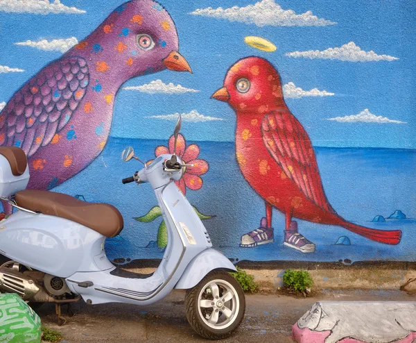 A Vespa scooter parked before a colorful street mural of two whimsical birds in Sète, a playful clash of everyday life and naive art