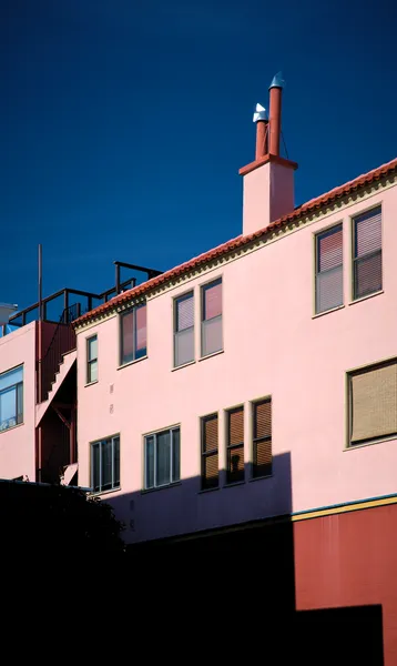 A pink-painted San Francisco house glows against a deep blue sky, its fire escape and chimney casting sharp shadows in this vivid color study of the city